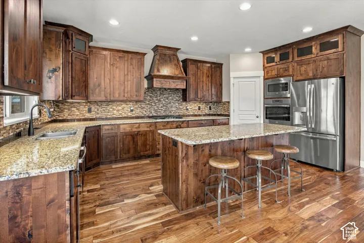 Kitchen featuring stained wood cabinetry, a central island with countertop seating, stone-tiled backsplash, custom range hood, and integrated stainless steel appliances