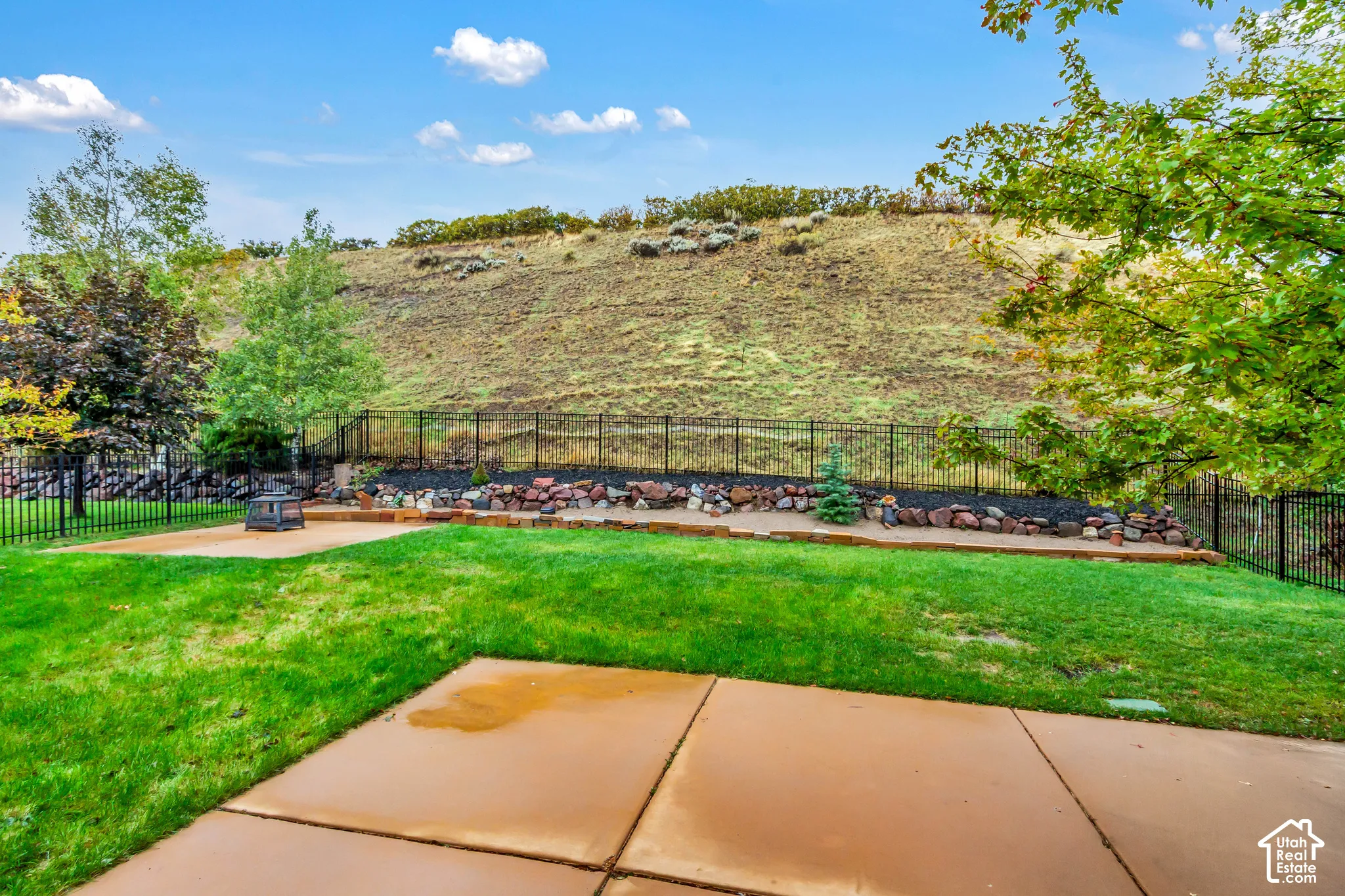 Rear yard featuring a concrete patio, manicured lawn, black metal fencing, and stone landscape terracing