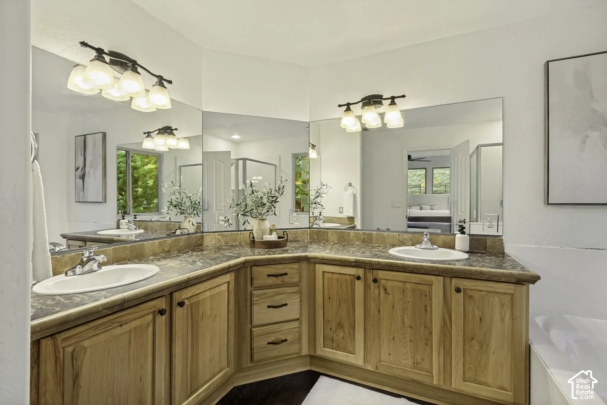 Corner vanity featuring wood cabinetry, natural stone-finish countertops, dual undermount sinks, and expansive multi-panel wall mirrors with multi-light fixtures