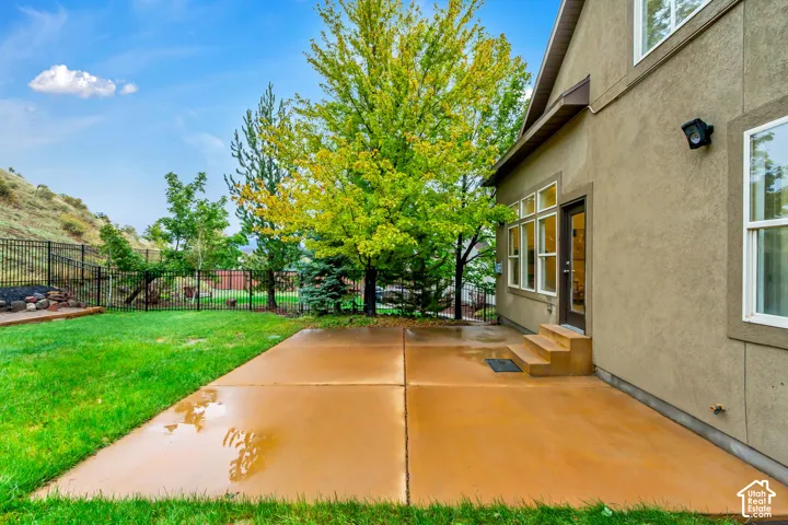 Rear exterior featuring a concrete patio, stucco siding, mature landscaping, and a metal perimeter fence