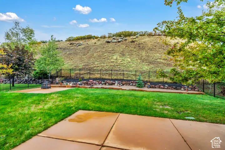 Rear yard featuring a concrete patio, manicured lawn, black metal fencing, and stone landscape terracing