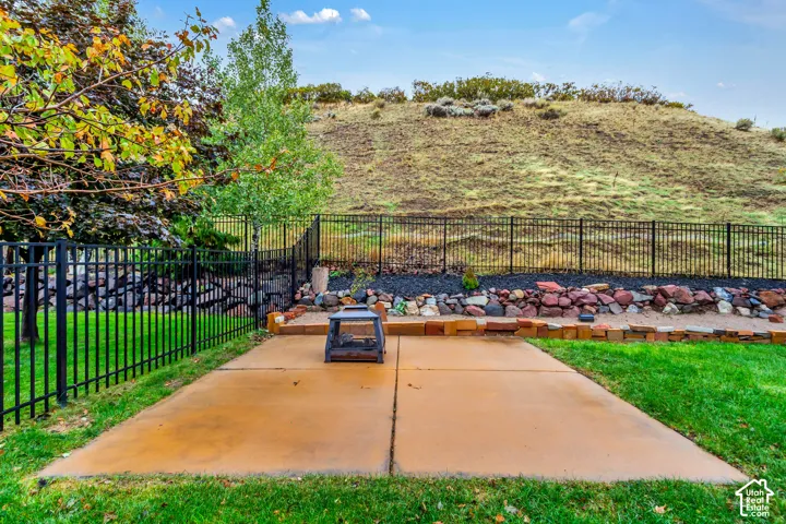 Backyard featuring a concrete patio, tiered stone retaining wall, black metal fencing, and mature trees against a natural hillside backdrop