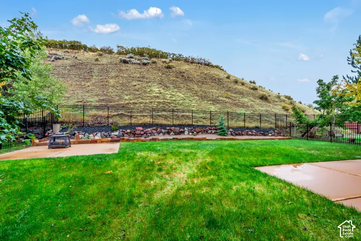 Backyard featuring a grass lawn, two concrete patios, a natural rock retaining wall, and a metal perimeter fence against a hillside backdrop