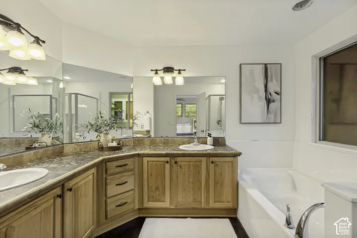 Extended vanity featuring wood-finish cabinetry, stone-texture countertop, dual integrated sinks, and multi-light wall fixtures, complemented by a deep soaking tub