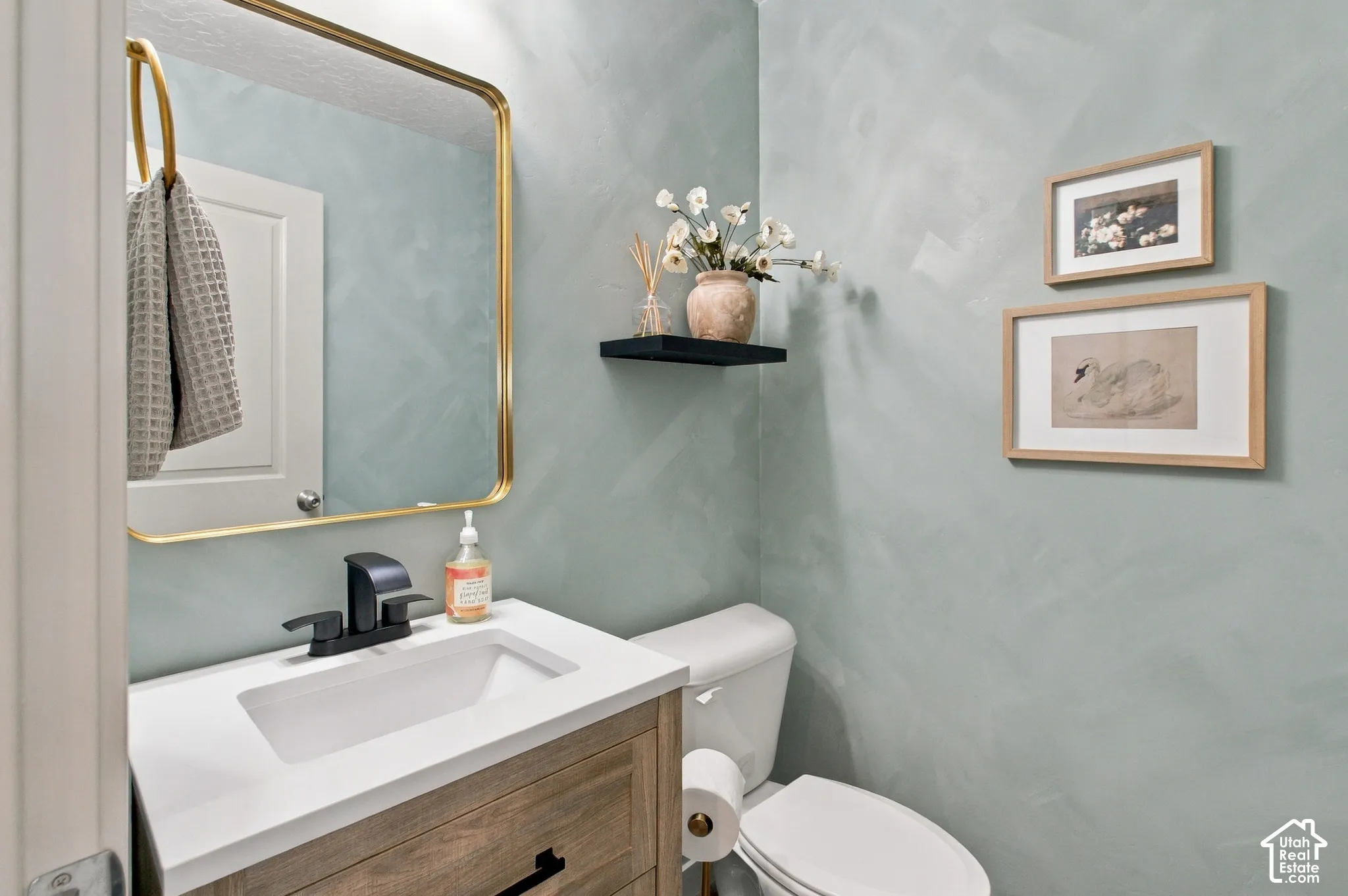 Textured light blue walls, wood-finish vanity with a white countertop, rectangular undermount sink, matte black faucet, and brass-framed mirror