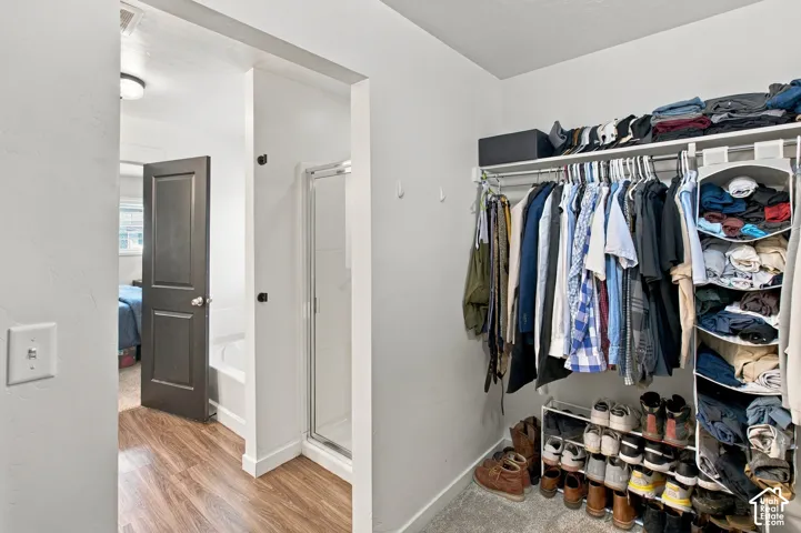 Walk-in closet featuring wire shelving and neutral carpeting, leading to a bathroom with a glass-enclosed shower, deep soaking tub, and wood-finish flooring