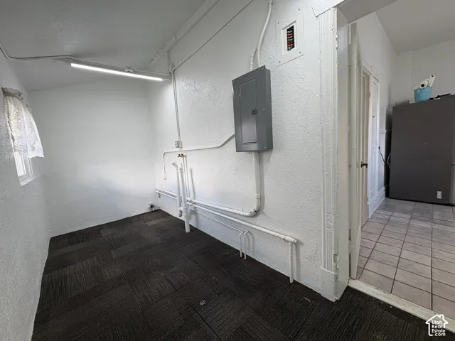 Utility space featuring dark tiled flooring, linear overhead lighting, exposed plumbing connections, and a mounted electrical panel