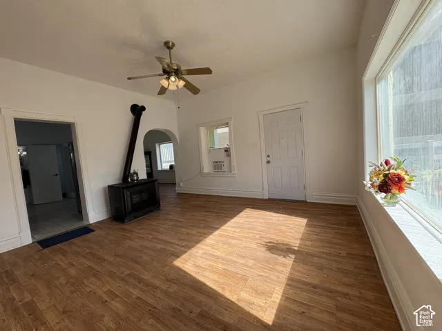 Wood-finish flooring, cast-iron style wood-burning stove with integrated flue, ceiling fan with multi-light fixture, expansive picture window, and arched doorway
