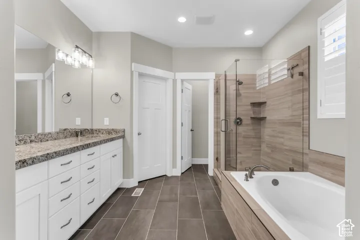 White cabinetry with stone-finish countertop, dual-sink vanity, glass-enclosed walk-in shower with wood-finish tile surround, separate soaking tub, and tile flooring