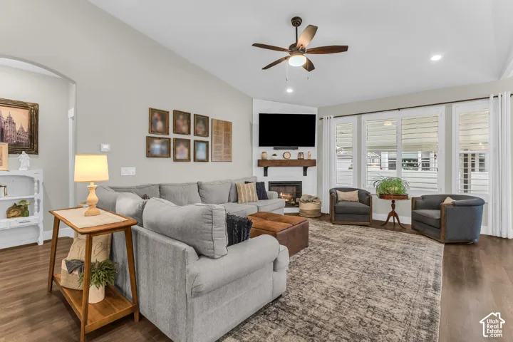 Vaulted ceiling living area featuring wood-finish flooring, corner fireplace with a timber mantel, and windows fitted with plantation shutters