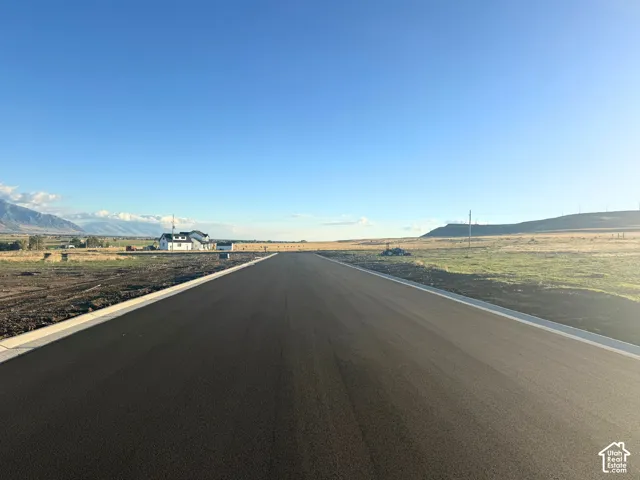 Newly paved asphalt road with concrete curbing, bordered by expansive open acreage and mountain horizons