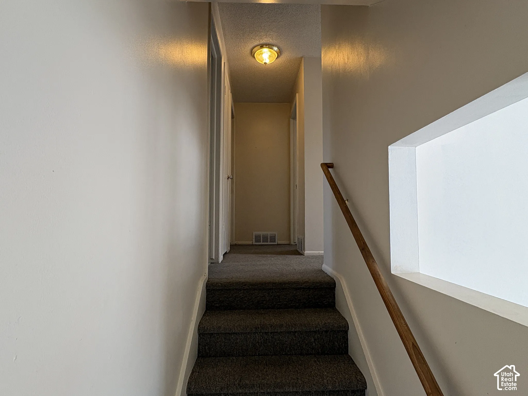 Stairway featuring neutral carpeted steps, a polished wood handrail, and a rectangular architectural cut-out window