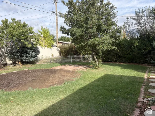 Backyard featuring a grass lawn, mature trees, a brick patio, a white metal fence, and stone paver edging
