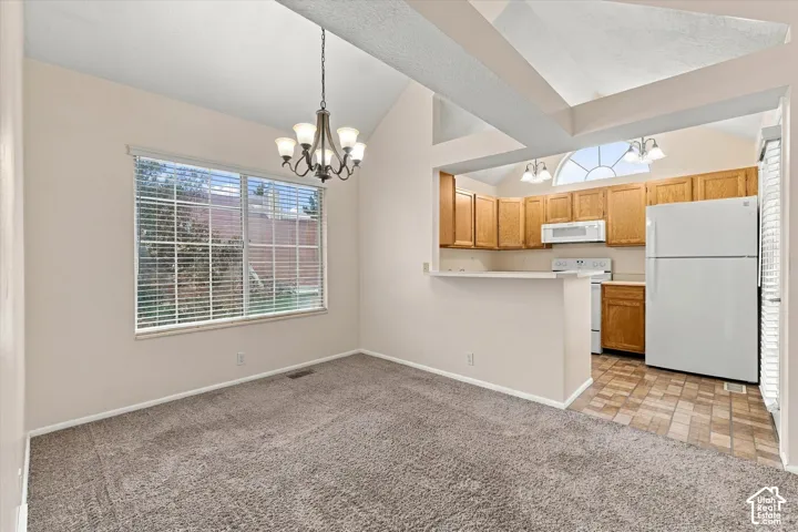 Vaulted ceilings featuring exposed beams, neutral carpet flooring, a multi-light chandelier, and a breakfast bar opening to a kitchen equipped with natural wood cabinetry