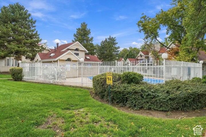 Community swimming pool enclosed by white metal fencing, surrounded by a manicured lawn, mature landscaping, and multi-story structures with pitched roofs