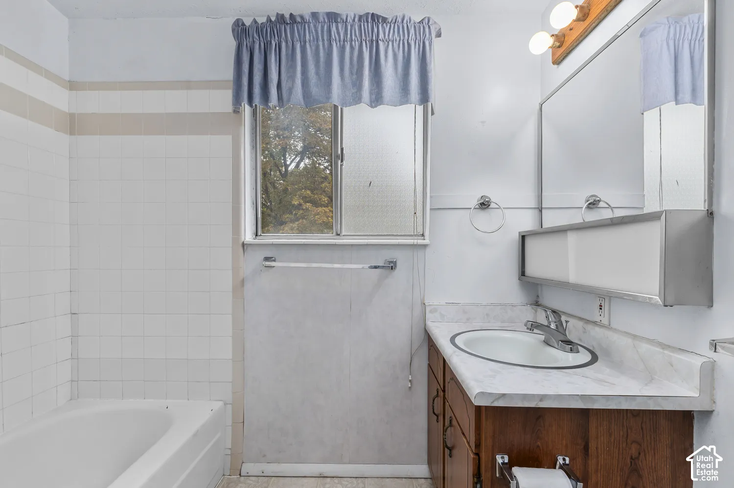 Bathroom featuring a white ceramic tile tub surround, sliding glass window, vanity with stone-patterned countertop, integrated sink with chrome faucet, and a large wall-mounted mirror