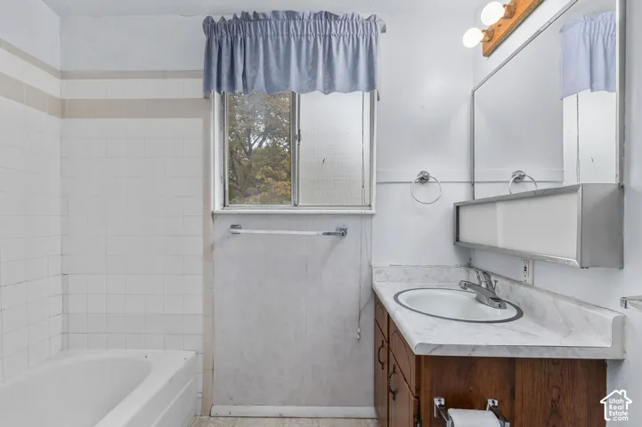 Bathroom featuring a white ceramic tile tub surround, sliding glass window, vanity with stone-patterned countertop, integrated sink with chrome faucet, and a large wall-mounted mirror