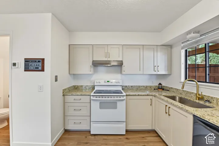 Kitchen featuring light-toned shaker cabinetry, stone-finish countertops, stainless steel range hood, undermount sink with a brass-finish faucet, and wood-finish flooring