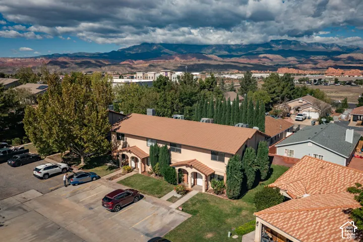 Multi-unit structure with earth-toned stucco exterior, shingled roofing, arched entryways, and manicured landscaping featuring mature trees, set against a backdrop of mountain ranges