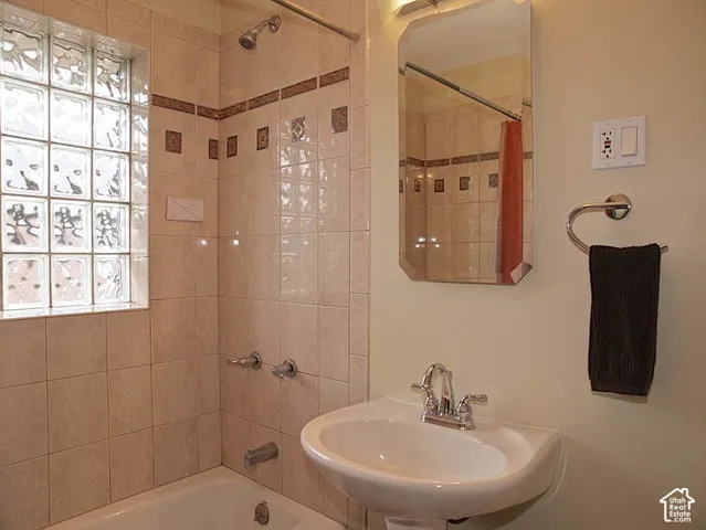 Bathroom featuring a glass block window, ceramic tile shower surround with decorative accent inlay, wall-mounted pedestal sink, polished chrome plumbing fixtures, and a frameless beveled edge mirror