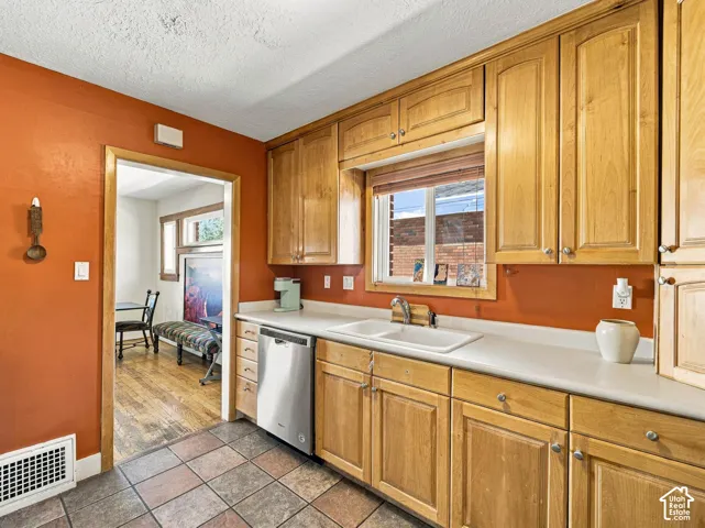 Kitchen featuring natural wood cabinetry, solid-surface countertops, dual-basin white sink, stainless steel dishwasher, and ceramic tile flooring