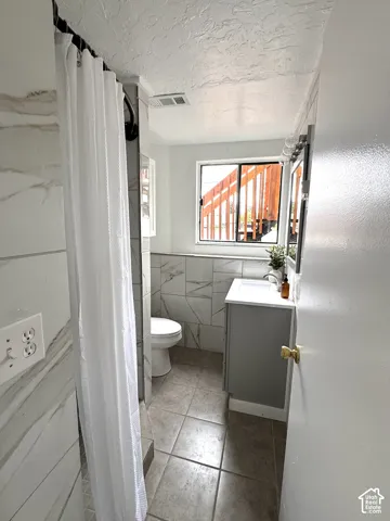 Bathroom featuring ceramic tile flooring, light-toned stone-patterned wall tiling, a modern grey vanity, and a window providing natural light