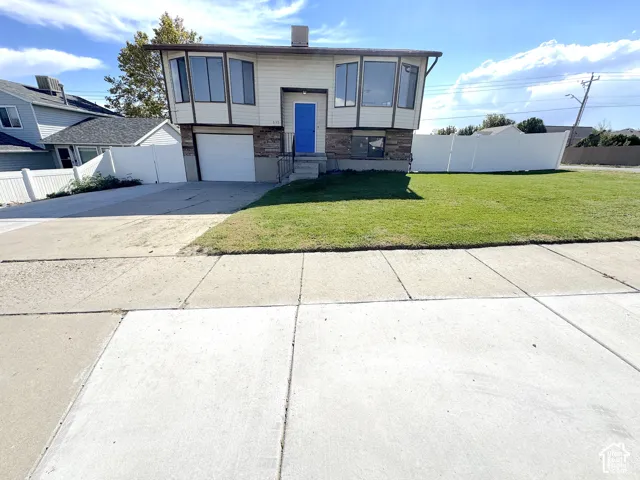 Split-level exterior featuring light-toned horizontal siding, stone veneer accents, multi-pane bay windows, a concrete driveway, and white vinyl perimeter fencing