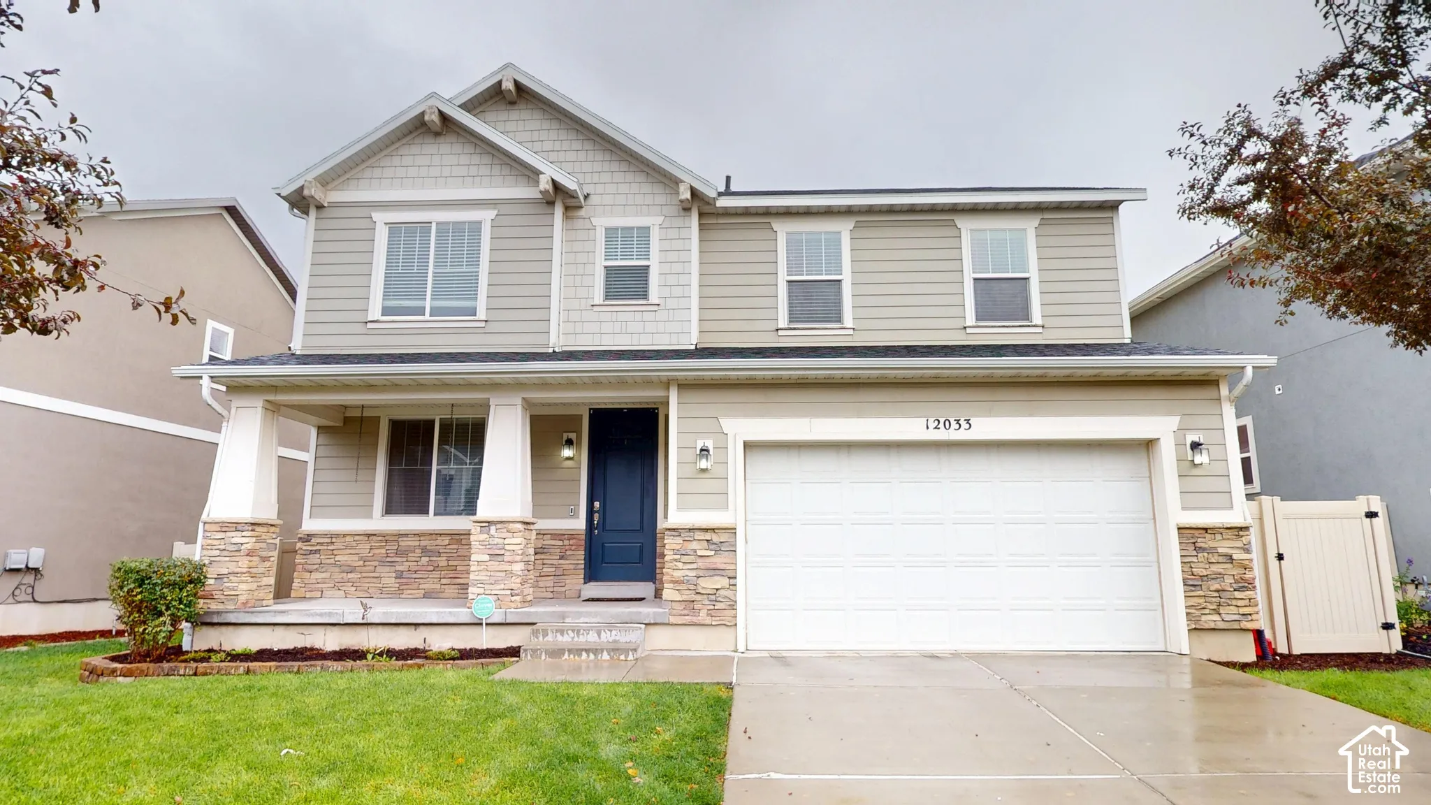Two-story exterior featuring tan horizontal siding, stone veneer accents, a covered front porch with tapered columns, and a two-car garage