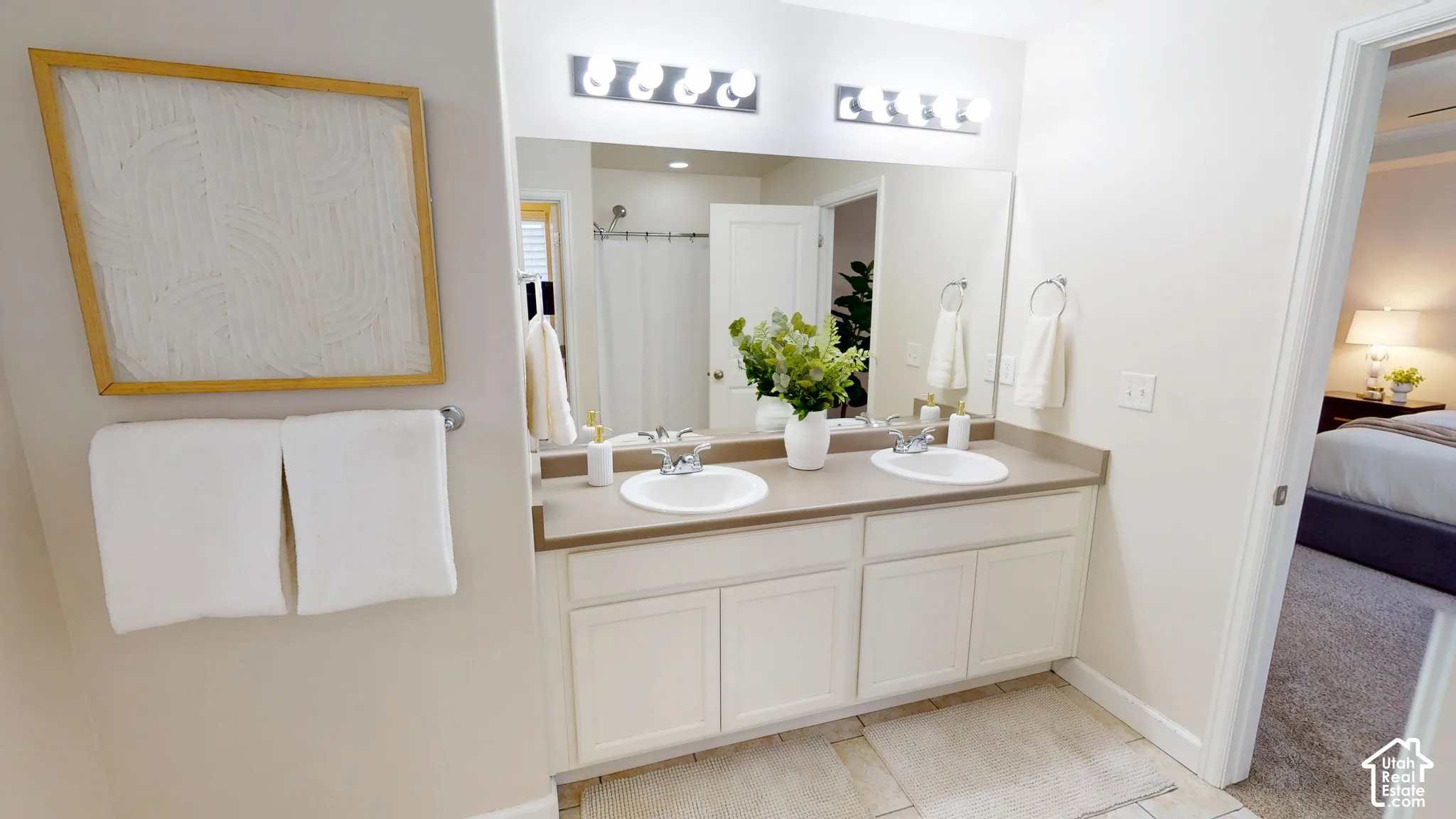 Primary suite bathroom with dual-sink vanity with a solid-surface countertop, white shaker-style cabinetry, and polished chrome fixtures, complemented by a large full-width mirror and multi-bulb wall lighting