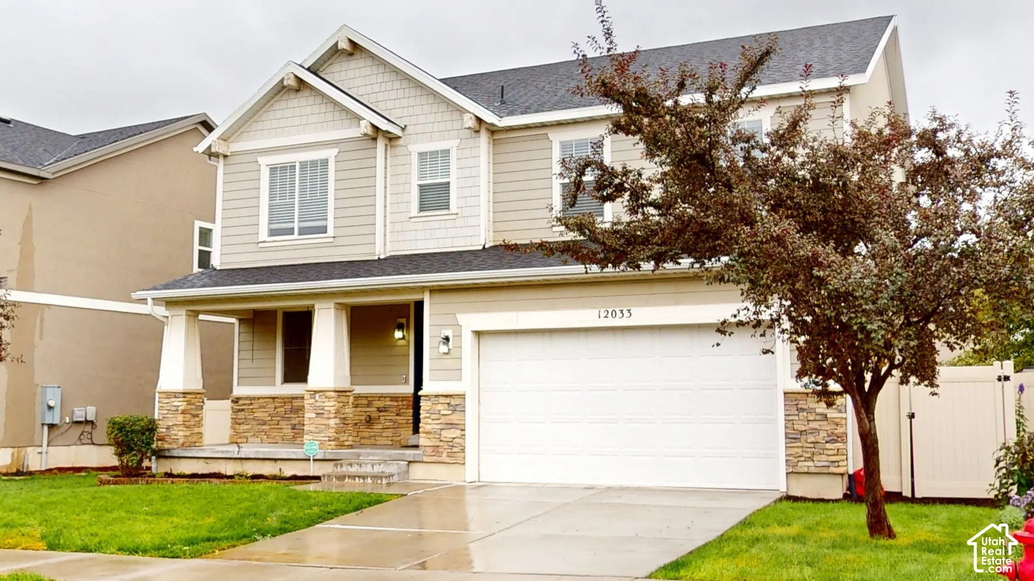 Two-story exterior featuring tan horizontal siding, stone veneer accents, a covered front porch with tapered columns, and a two-car garage