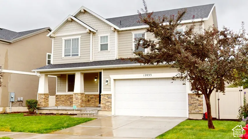 Two-story exterior featuring tan horizontal siding, stone veneer accents, a covered front porch with tapered columns, and a two-car garage