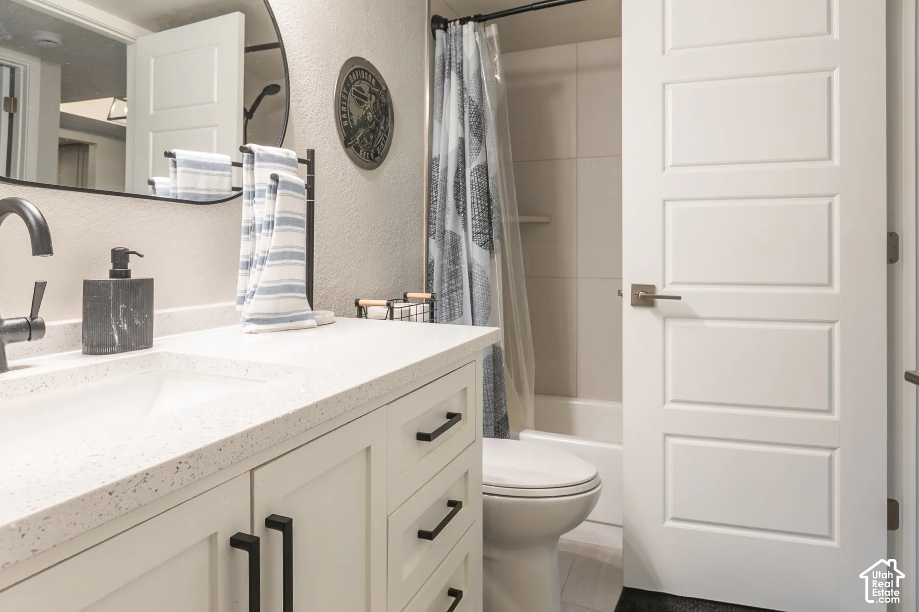 Bathroom featuring a vanity with a light-toned countertop, integrated rectangular sink, matte black hardware, a circular wall-mounted mirror, and a tub-shower combination with light-toned wall tile