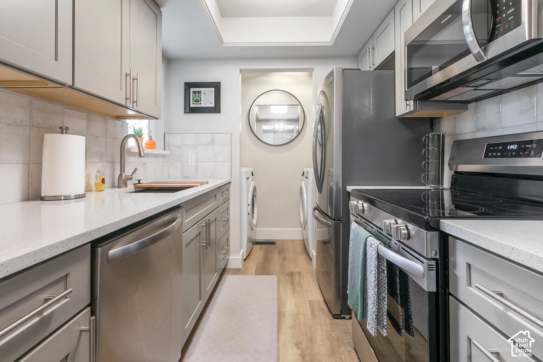 Galley-style kitchen featuring light-gray shaker cabinetry, quartz countertops, tile backsplash, stainless steel appliances, and wood-finish flooring