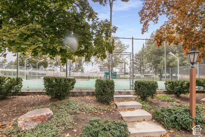 Tennis court enclosed by chain-link fencing, featuring concrete stairs, manicured evergreen shrubs, and built-in lantern-style lighting