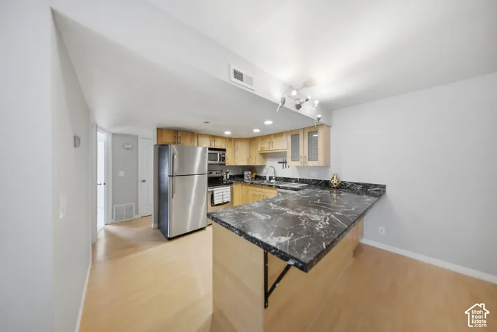 Kitchen featuring wood-finish flooring, stainless steel appliances, light wood cabinetry, a dark stone countertop, and a breakfast bar