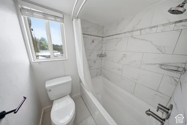 Bathroom featuring a white bathtub, shower with white and grey variegated tile surround, brushed nickel fixtures, a white toilet, and a window with blinds