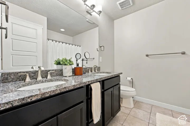 Dual-sink vanity with stone-finish countertop, dark cabinetry, full-width vanity mirror, tile flooring, and brushed-nickel hardware