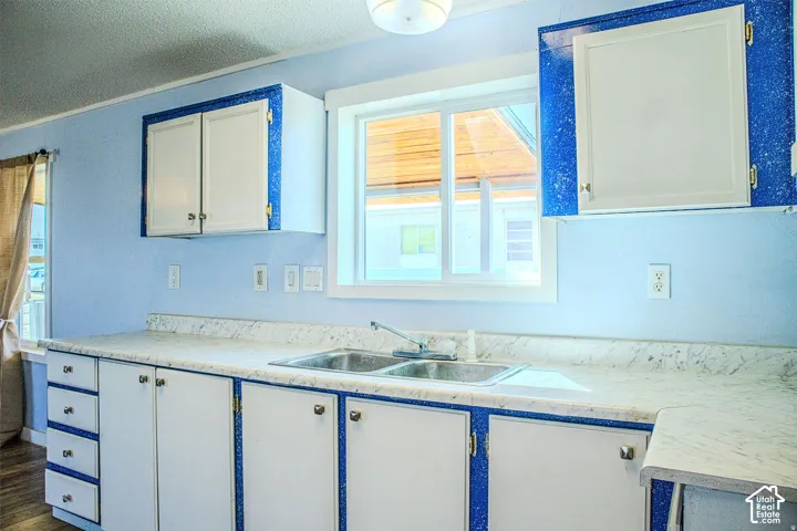 Kitchen featuring white cabinetry with blue accents, light-toned stone-patterned laminate countertops, dual-basin stainless steel sink, and a dual-pane sliding window