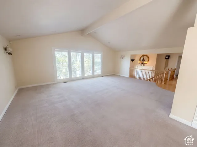 Vaulted ceiling featuring exposed beams, neutral carpeting, large windows with interior shutters, and wooden spindle railing
