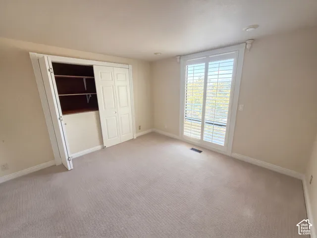 Neutral-toned interior space featuring carpeted flooring, bi-fold closet doors with built-in shelving, and a large window accented with white plantation shutters