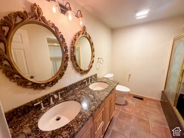 Dual-sink vanity with natural stone countertop, light wood cabinetry, integrated oval basins, and metallic-finish faucets, complemented by terra-cotta tile flooring and a glass-enclosed shower