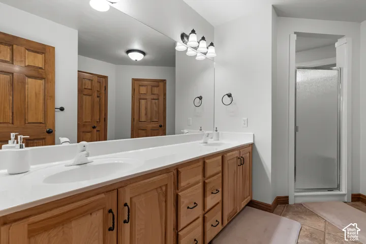 Dual-sink vanity with a solid-surface countertop, full-width wall mirror, natural wood-finish cabinetry, tile flooring, and a walk-in shower enclosure
