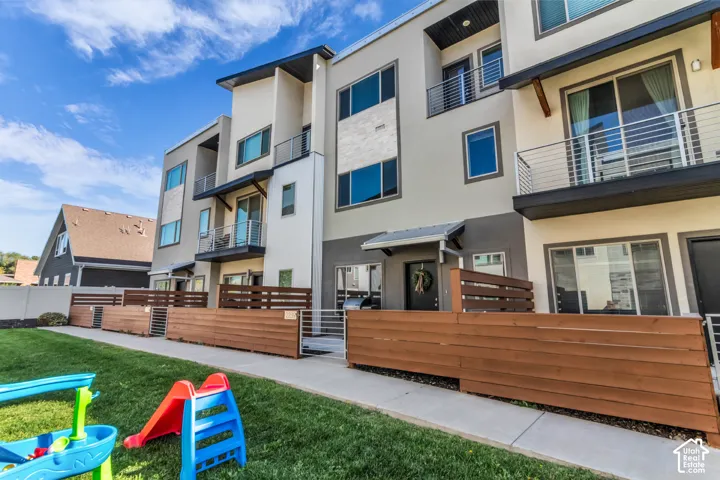 Modern three-story townhouse exterior featuring a neutral stucco finish, stone veneer accents, wood-slat privacy fencing, multiple private balconies with metal railings, and concrete walkways