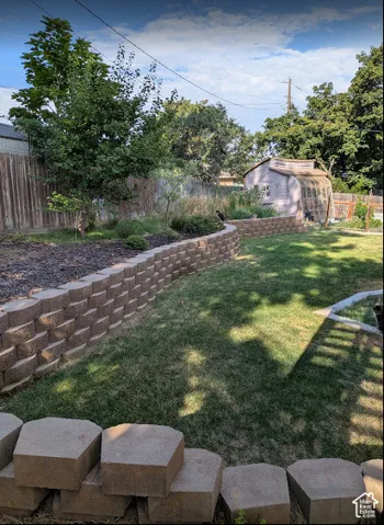 Backyard featuring tiered segmental block retaining walls, mature trees, green lawn, and a detached shed