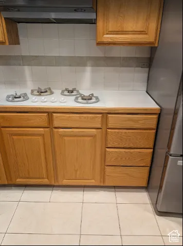 Kitchen featuring light wood-finish cabinetry, solid surface countertop, four-burner gas cooktop, stainless steel ventilation hood, and tile backsplash