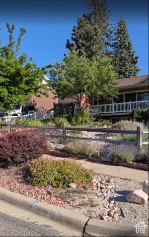 Landscaped front yard featuring a concrete walkway, tiered rock garden, split-rail wooden fencing, mature trees, and a white deck railing