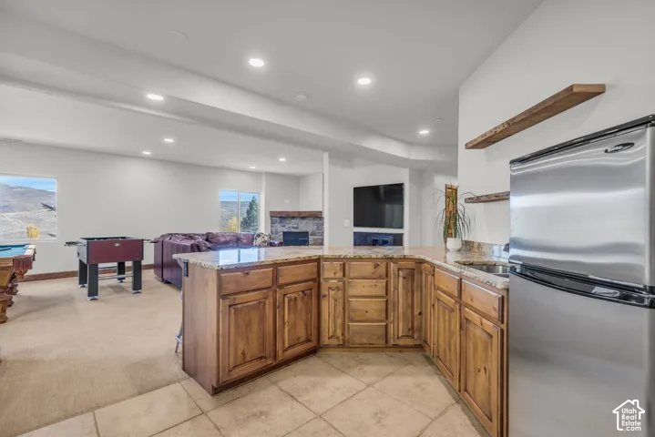 Full lower-level entertainment space featuring an integrated kitchenette with wood cabinetry, light-toned countertops, and a stainless steel refrigerator