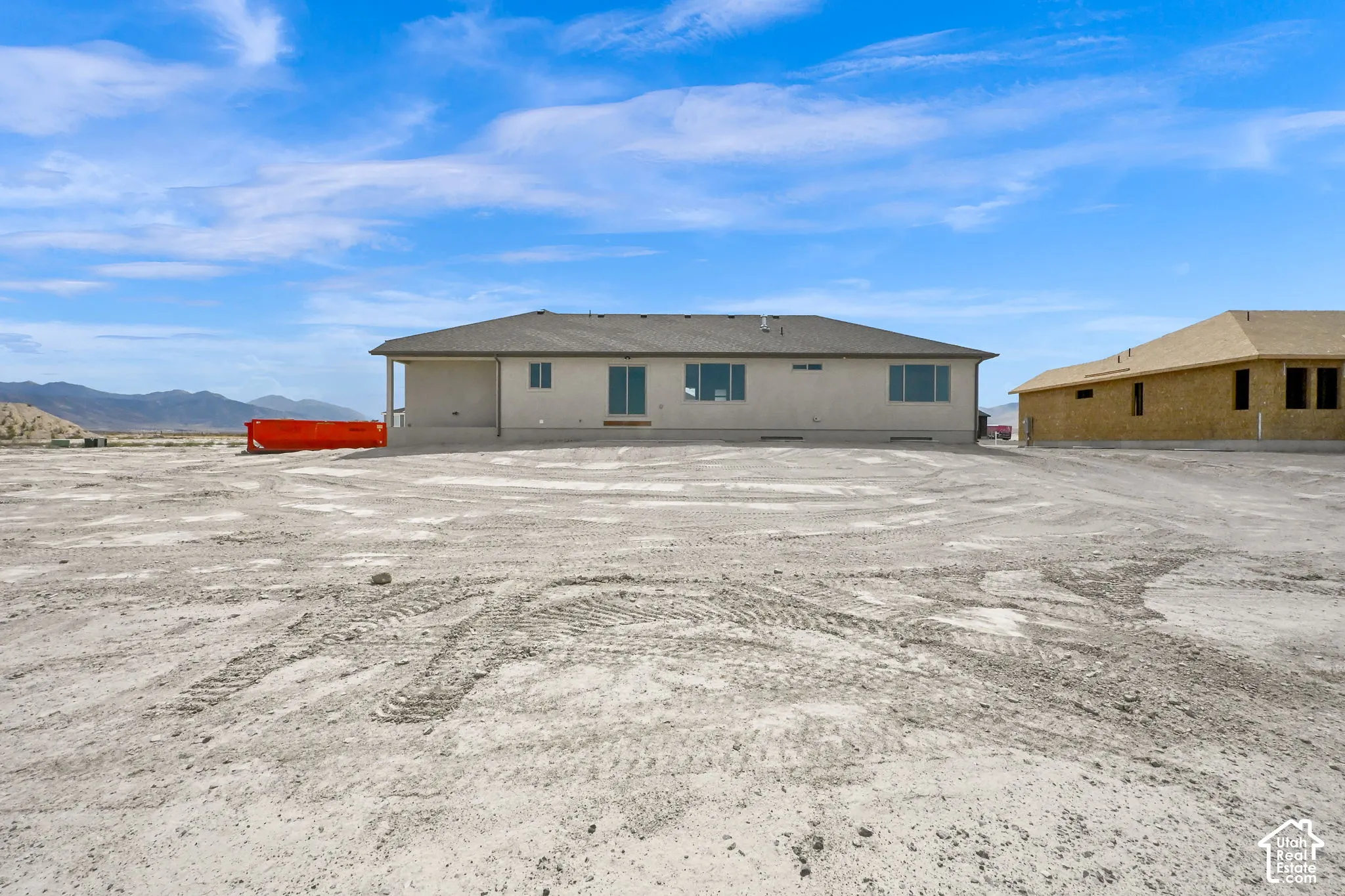Single-story exterior featuring light-toned stucco siding, a hip roof with asphalt shingles, sliding glass doors, and expansive lot space with mountain backdrops