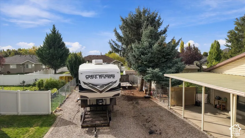 Expansive gravel side yard featuring a covered patio, vinyl perimeter fencing, mature evergreen trees, and specialized reinforced pad for large-vehicle storage