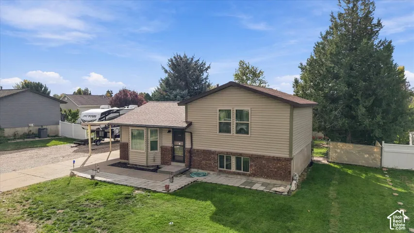 Split-level exterior featuring tan vinyl siding, a brick masonry foundation, asphalt shingle roof, stone-paver patio, and attached carport